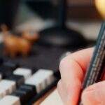 Hand markiert Aufgaben in einem Notizbuch neben einer Tastatur als Symbol für dokumentierte KI-Kompetenz und Betriebsleitplanken