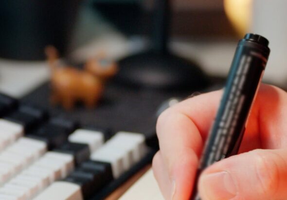 Hand markiert Aufgaben in einem Notizbuch neben einer Tastatur als Symbol für dokumentierte KI-Kompetenz und Betriebsleitplanken